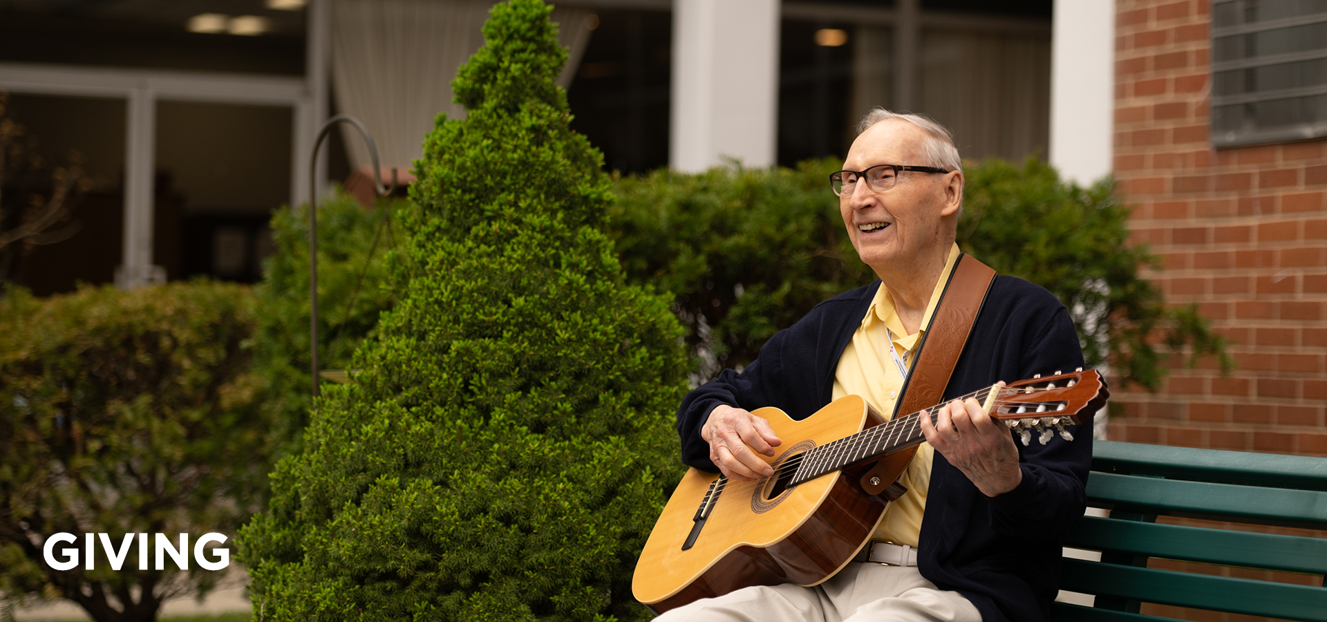 UMCF giivng new HEADER An older gentleman smiles as he strums his guitar on a bench in the Pitman Manor courtyard.