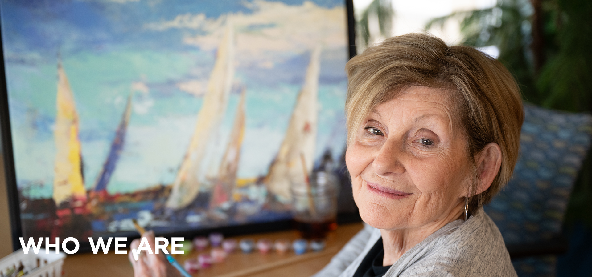 UMCF who we areHEADER V3 An artistic resident of Collingswood Manor smiles as she sits in front of a painting of sailboats. She holds a paintbrush and has paints arranged on her table.