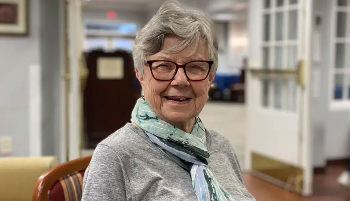 bristol-glen A Bristol Glen resident wearing glasses and a scarf smiles in the hallway of her senior living community.