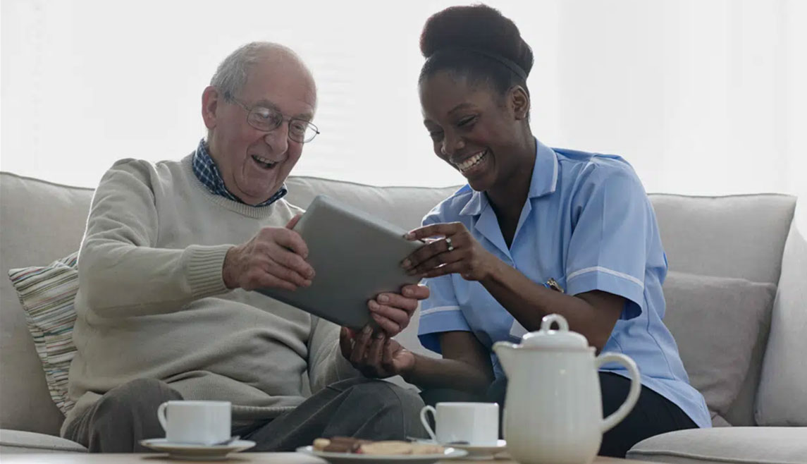 A smiling, uniformed home healthcare worker helps her elderly male client use a tablet. This is a stock image.