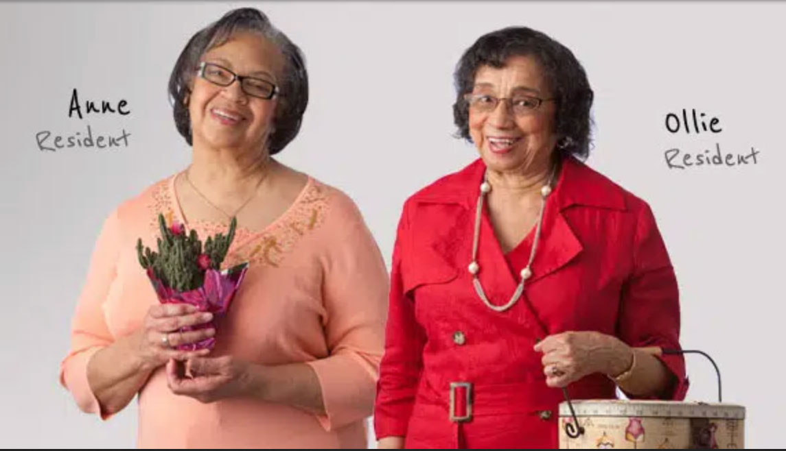 Two older women pose for a photo shoot at their UMC senior living community. The woman on the left wears a peach sweater and holds a potted plant, while the woman on the right wears a red suit and carries a purse.