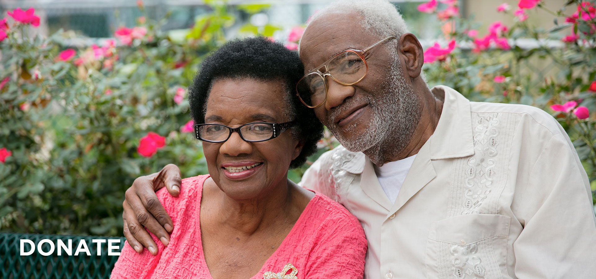 donate-header-couple A couple in their eighties smile as they sit on a bench in the garden of a UMC senior affordable housing community. Both the man and woman are wearing glasses and there are pink flowering bushes behind them.