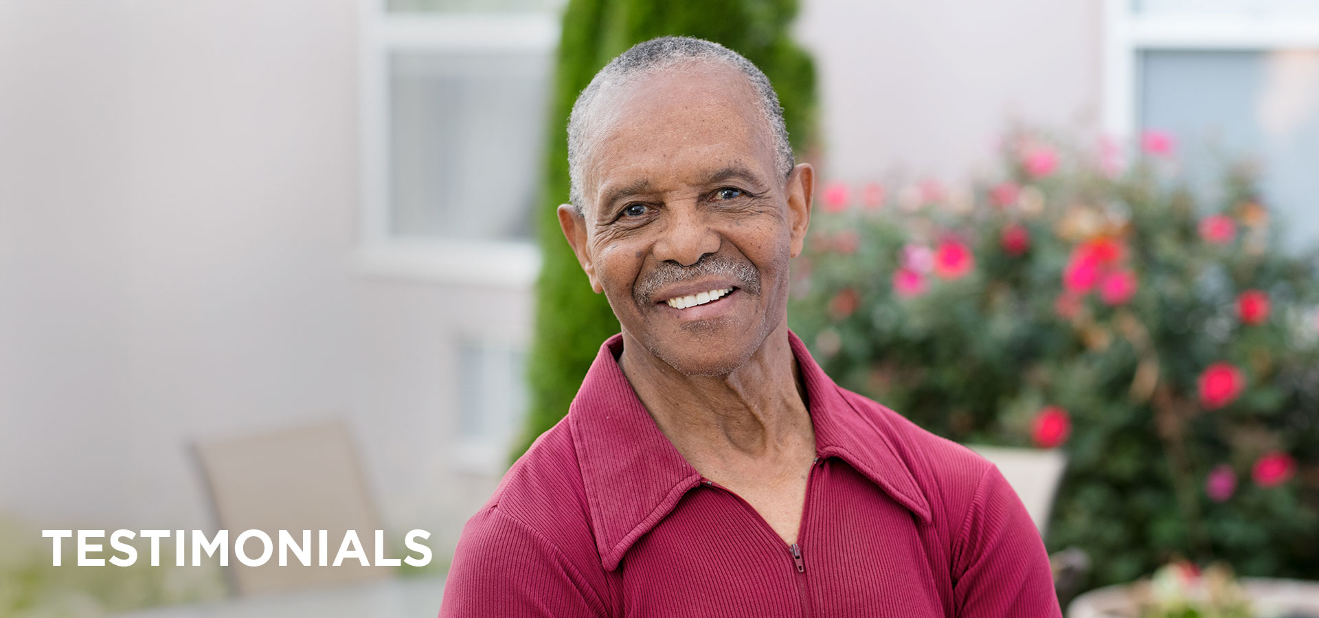 testimonials-header A resident wearing a maroon polo shirt smiles in front of a patio table, chairs & rose bush outside his senior living community.