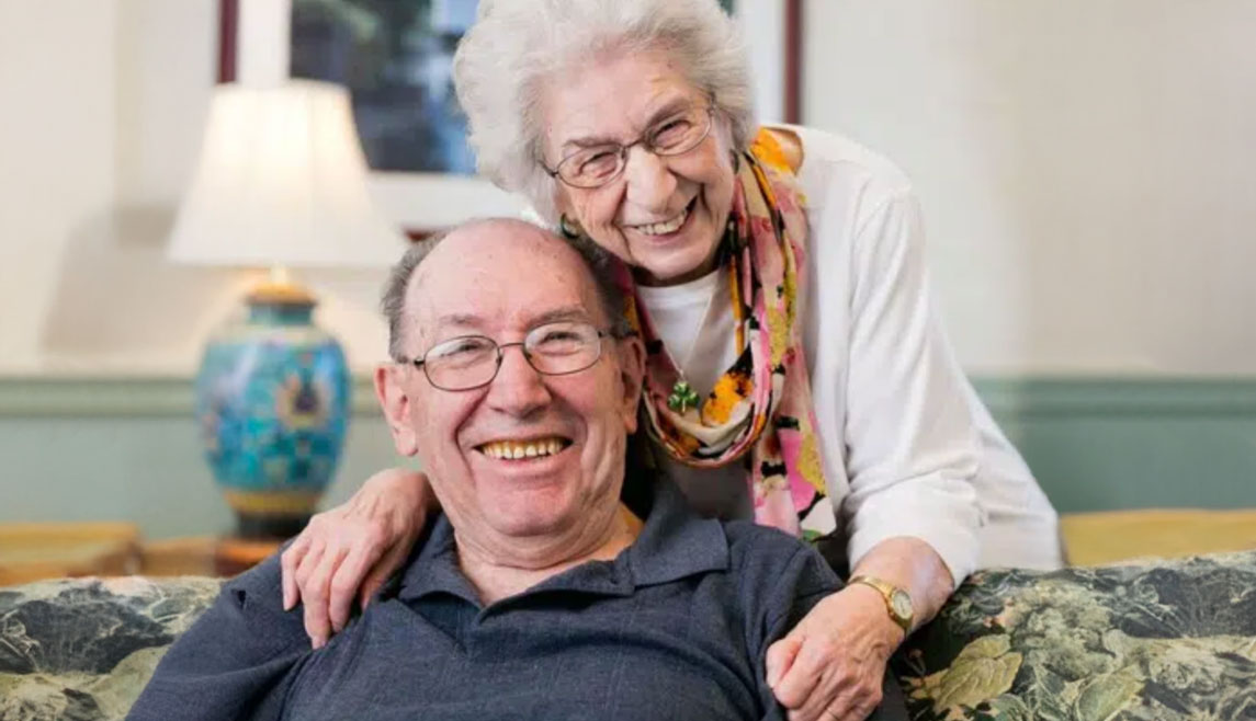 An older couple smile in their apartment at a UMC senior living community. The man is seated while the woman stands behind him with her arms around his shoulders.